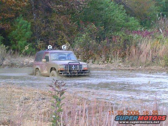 Muddy 1989 Ford Bronco II driving through a muddy puddle with trees in the background