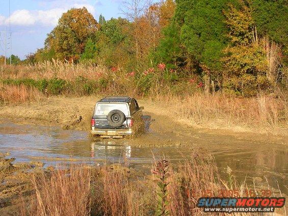 1989 Ford Bronco II driving through a muddy puddle with tall dry grass and trees in the background