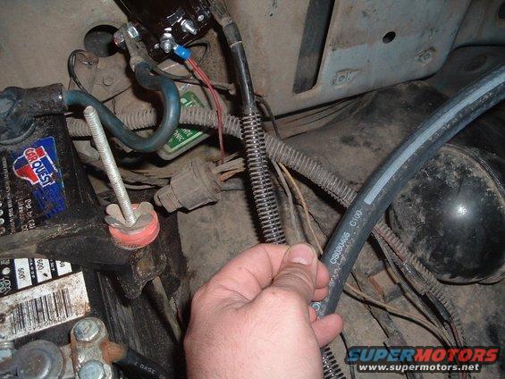 Close-up of a vehicle's engine bay showing the battery, wiring, and a hand holding a hose