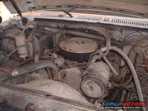 Engine bay of a white 1980s Ford truck with a V8 engine, air filter, and hoses. The engine is dusty and appears to be in a used condition