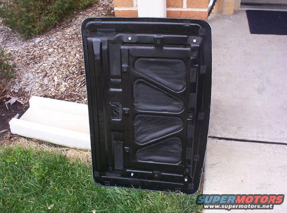 Underside of a black car hood, showing structural ribs and insulation, outdoors on grass and concrete