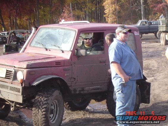 Maroon Suzuki Samurai with people inside, parked in mud. Another truck is visible in the background