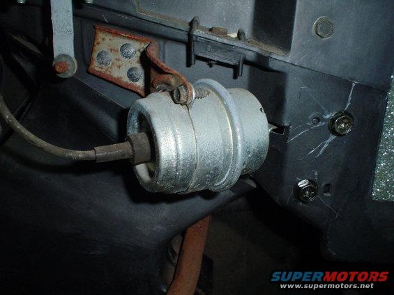 Close-up of a silver cylindrical component with a cable attached, mounted to a black plastic panel with a rusty bracket