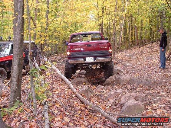 Red Dodge pickup truck with 4x4 badging navigating a rocky forest trail. Another off-road vehicle is visible to the left
