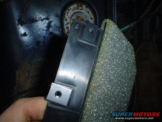 Close-up of a black plastic car part with metal brackets, next to foam padding. A finger is visible
