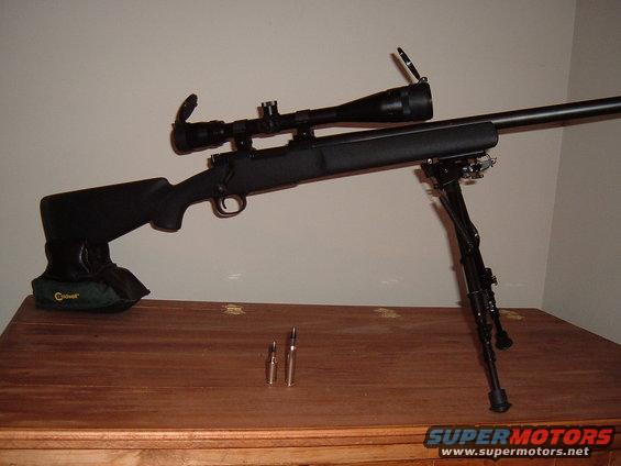 Black rifle with scope and bipod on a wooden surface, with two bullets in front