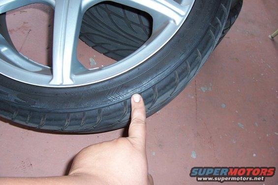 Close-up of a tire on a silver alloy wheel. Finger points to tread