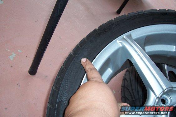 Close-up of a tire with a finger pointing to the sidewall. A silver alloy wheel is visible. A black metal leg is in the background