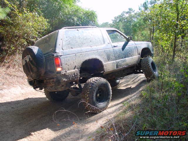Muddy SUV with large tires driving on a dirt trail in a wooded area. Spare tire mounted on the back
