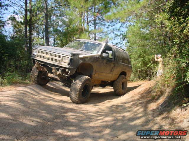 Muddy brown Chevrolet Blazer K5 off-roading on a dirt path, front-left tire angled up