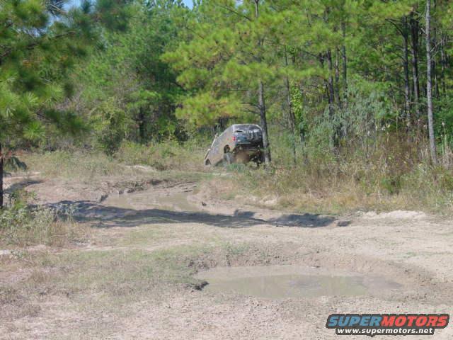 Rear view of a light-colored SUV navigating a muddy trail through trees
