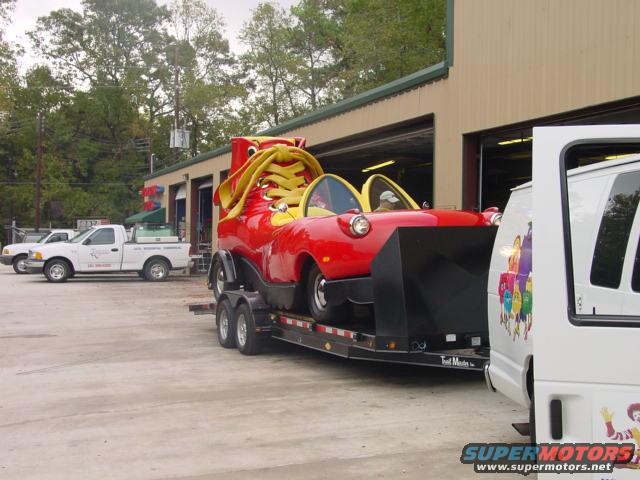 Red novelty shoe car on a trailer, with a white van and two pickup trucks nearby