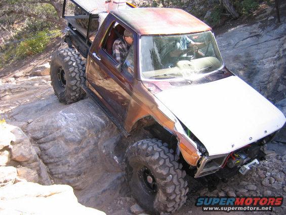 Modified pickup truck with large tires navigating rocky terrain. White hood, brown body, winch visible