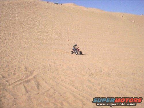 ATV rider on a sand dune, with other people and vehicles visible further up the dune