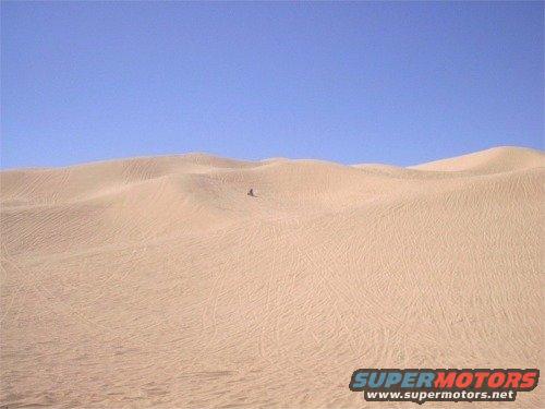 Sand dunes under a clear blue sky with a small ATV visible in the distance