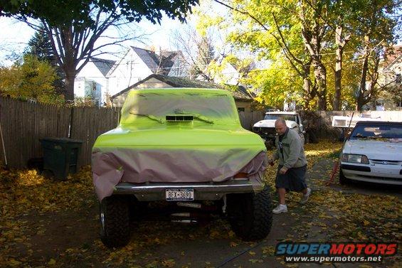 Rear view of a Bronco covered in green plastic, with a man nearby and another car parked to the right. License plate reads BEX-3690