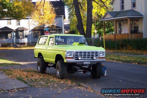Lifted lime green SUV with a hood scoop parked on a street with fallen leaves. A person is working underneath it