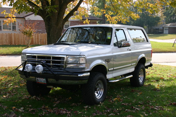 Silver Ford Bronco parked on grass with a tree and house in background. Has a brush guard with lights