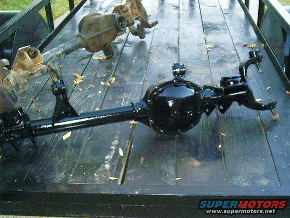 Two vehicle axles, one black and shiny, the other rusty, sit on a wooden trailer bed