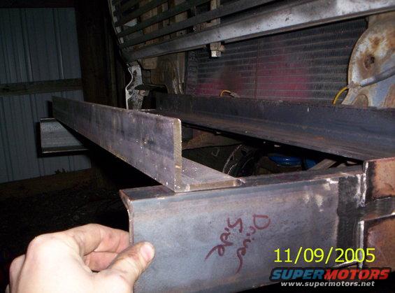 Metal frame pieces attached to a vehicle's front end, with a radiator visible behind. A hand is holding a piece of metal