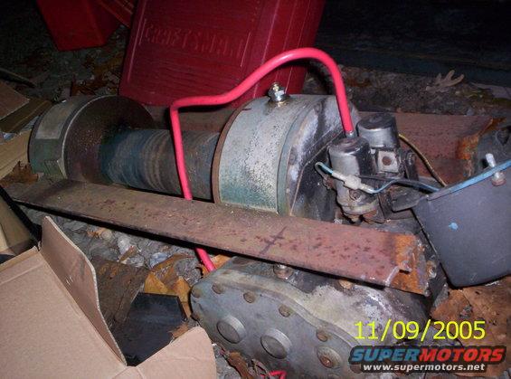 Winch mechanism with red hoses and electrical components, resting on rusty metal