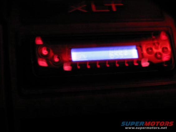 Car stereo with glowing blue display and red buttons, above it is an illuminated XLT badge