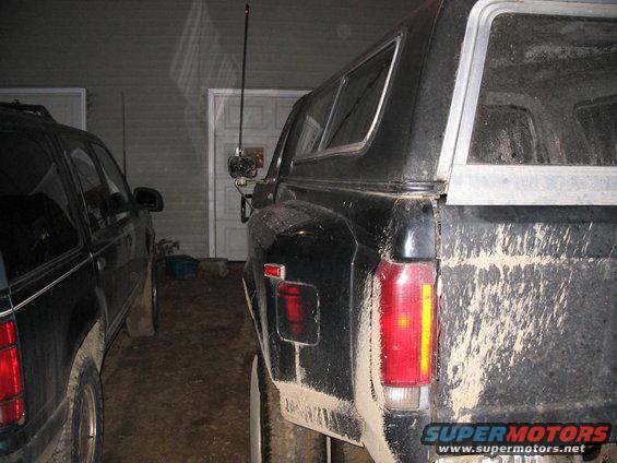 Rear quarter view of a black pickup truck with a camper shell, covered in mud. Another dark SUV is parked next to it