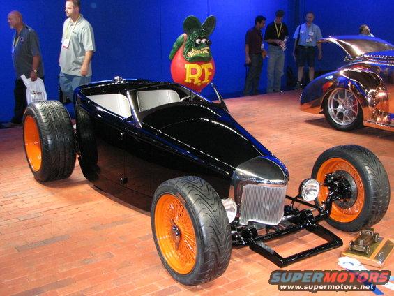 Black hot rod with orange wire wheels and exposed front suspension, seen from a 3/4 front angle