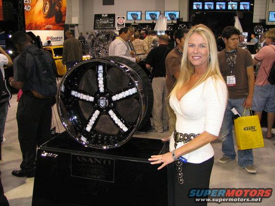 Woman posing next to a large custom wheel with diamond-like accents. The wheel has 'ASANTI' written on its stand