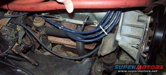 Engine bay with blue spark plug wires, red hose, and metal components. A hand is visible at the top
