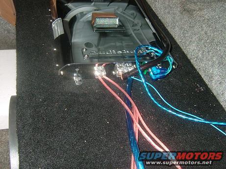 Car amplifier with pink and blue wires connected to terminals, mounted on a carpeted surface