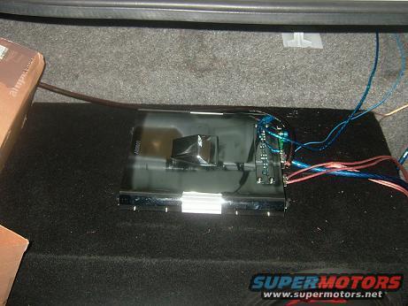 Car amplifier with blue and red wires connected, sitting on a black subwoofer box in a car trunk