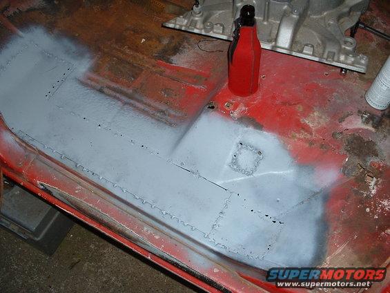 Car floor pan with gray primer applied over patched areas. Red metal visible around edges