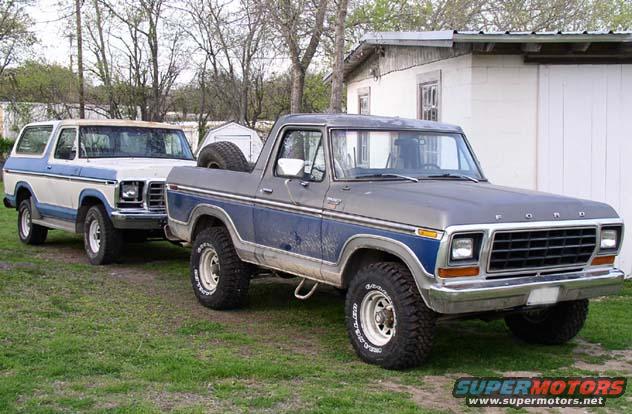 Two Ford Broncos parked on grass in front of a building. The front Bronco is gray and blue, the rear one is white and blue