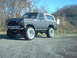 1986 Ford Bronco - This lifted Ford Bronco has a white removable top and is parked on a gravel surface with trees in the background.