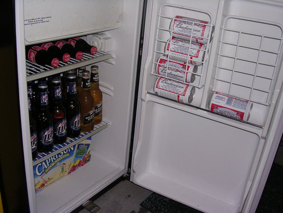 Open refrigerator interior with shelves stocked with Budweiser cans, Miller Lite bottles, Coca-Cola bottles, and a Capri Sun juice box