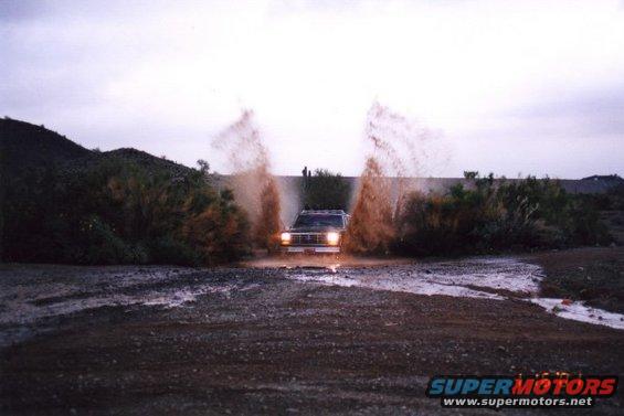 Dark SUV driving through water, kicking up large plumes of brown spray