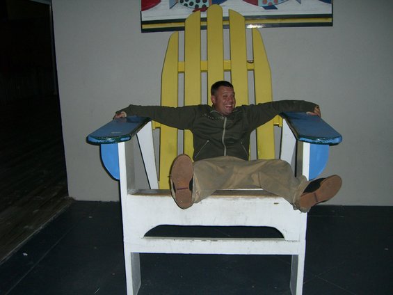 Man with arms outstretched sits in a giant yellow and white Adirondack chair with blue armrests