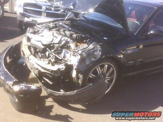Front end of a black car severely damaged, hood open. Another vehicle is visible behind it