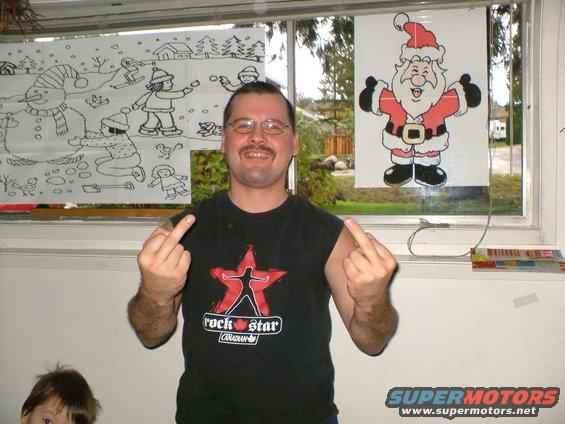 Man in a black t-shirt with a star logo gives the middle finger to the camera, with holiday drawings on the window behind him