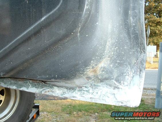 Close-up of a vehicle's lower body panel showing rust and repair work. A tire is visible on the left