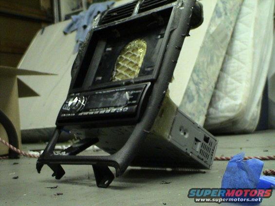 Car stereo and black dashboard trim piece with vents and a gold honeycomb pattern behind them. Some mattresses are in the background