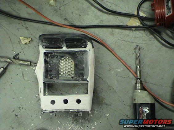 Car dashboard center console piece with air vents, mesh, and three circular cutouts, next to a drill