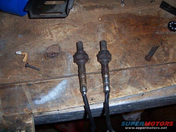 Two used oxygen sensors on a wooden workbench with a screw and other small items