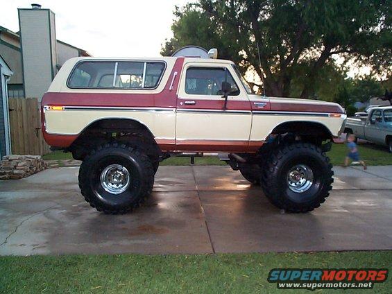 Lifted 1978 Ford Bronco with two-tone paint, large off-road tires, and a child running in the background near another pickup truck