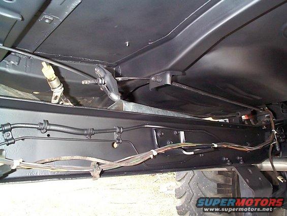 Underbody view of a vehicle, showing brake lines, wiring harness, and frame components. Tire visible at bottom right