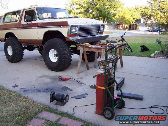 Lifted Ford Bronco on sawhorses, with welding equipment and metal pieces nearby