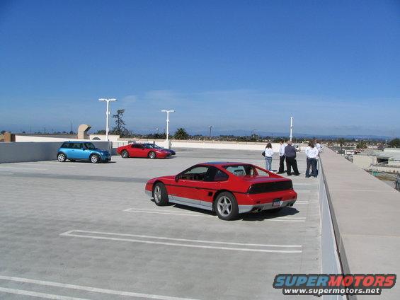 Red Pontiac Fiero parked on a rooftop lot with a blue Mini Cooper and another red sports car