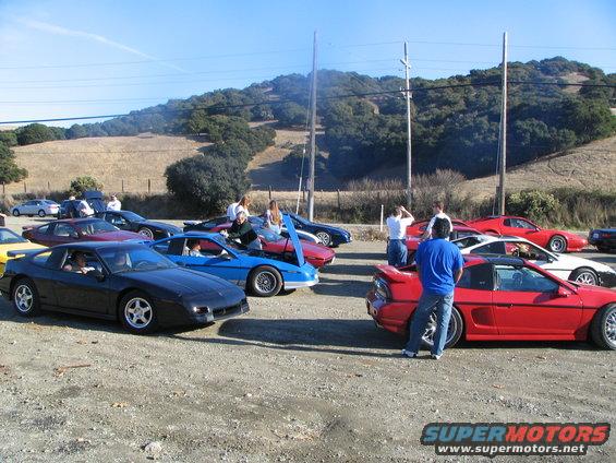 Several Pontiac Fieros parked together, one black, one blue, one red. Other sports cars also present