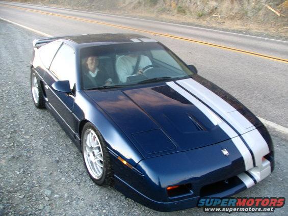 Blue 1986 Pontiac Fiero with silver racing stripes on the hood and roof, driving on a road